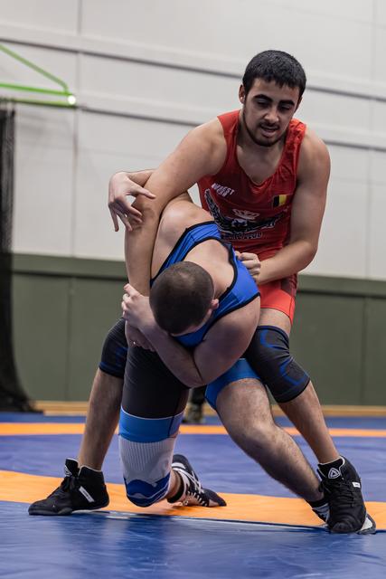 Young wrestler in red singlet dominates opponent in blue, gripping firmly on blue competition mat indoors.