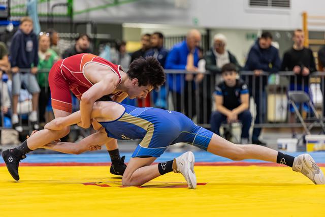 Wrestler in red singlet aggressively drives blue-clad opponent down onto the yellow mat during an intense match.