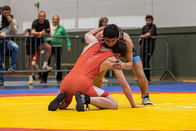 Two young wrestlers grapple intensely on a yellow mat, one in red kneeling as the other dominates from above.