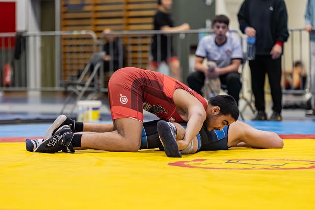 Wrestler in red singlet dominates opponent in blue, pressing them down on the yellow competition mat.