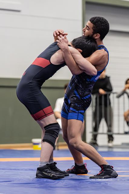 Two young wrestlers locked in a standing clinch on a blue mat, straining with intensity during an indoor match.