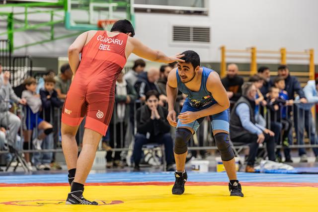 Two wrestlers in red and blue singlets face off on a yellow mat, crowd watching intently in the background.