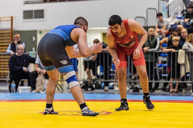 Two wrestlers in blue and red singlets square off on a yellow mat, both crouched and focused, crowd watching behind barriers.