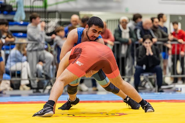 Young wrestler in blue singlet smiles while controlling opponent in red during match on yellow mat, crowd watching.