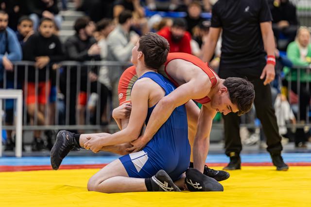 Wrestler in red lifts opponent in blue from behind on yellow mat, crowd watching in background.
