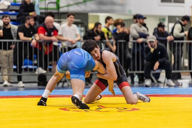 Two young wrestlers lock up on a yellow mat, one in blue singlet crouching low, the other in black staying balanced.