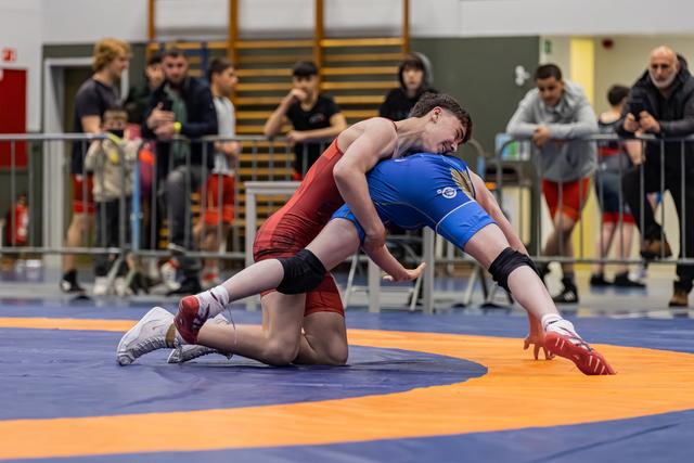 Two young wrestlers in red and blue singlets battle on the mat as spectators watch from behind barriers.