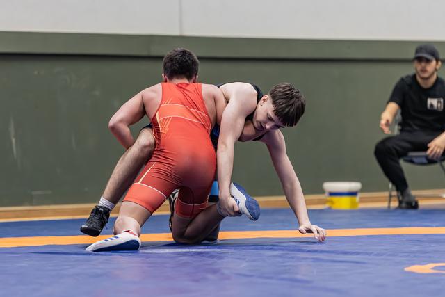 Teen wrestler in black singlet drives forward aggressively while opponent in red struggles on knees; referee observes from sideline.