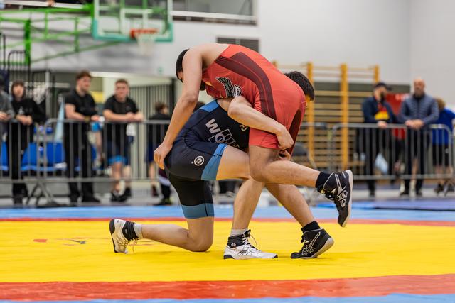 Two wrestlers battle for control, one in blue singlet on knee, other in red driving forward on colorful competition mat.