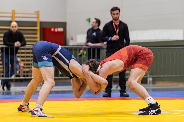 Two wrestlers in blue and red singlets locked in an intense clinch on a yellow mat, referee watching closely.