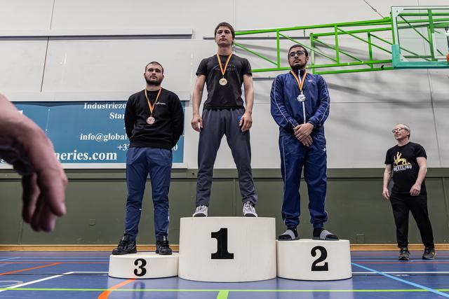 Three male wrestlers stand on a podium, gold medalist centered on platform 1, flanked by silver and bronze winners in a gym.