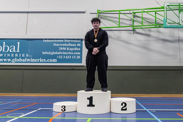 Young male athlete stands proudly on the first-place podium, wearing a gold medal, in an indoor sports hall.