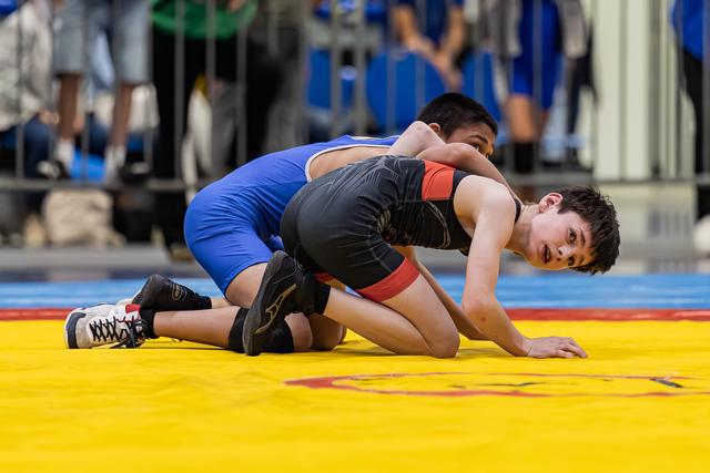 Young wrestler in black singlet controls opponent on yellow mat, focused expression during intense match
