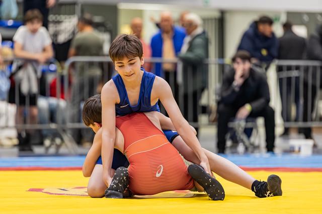 Boy in blue singlet dominates opponent in red, pinning him to the yellow mat with focused concentration.