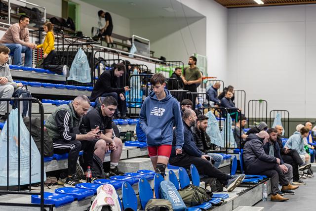 Teen in New Balance hoodie and red shorts walks pensively through blue stadium seats among waiting athletes.