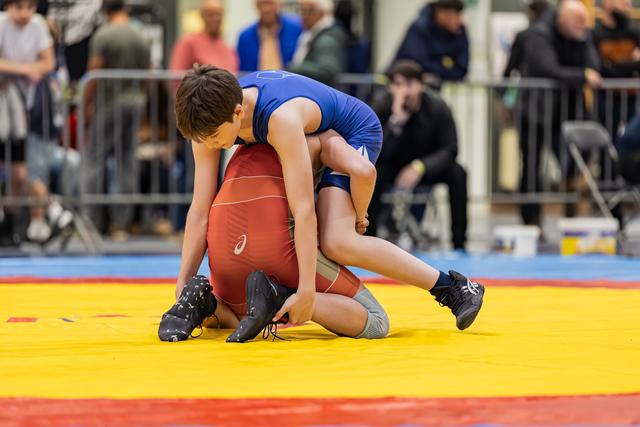 Young wrestler in blue singlet dominates opponent in red, gripping legs on a yellow mat with crowd watching.