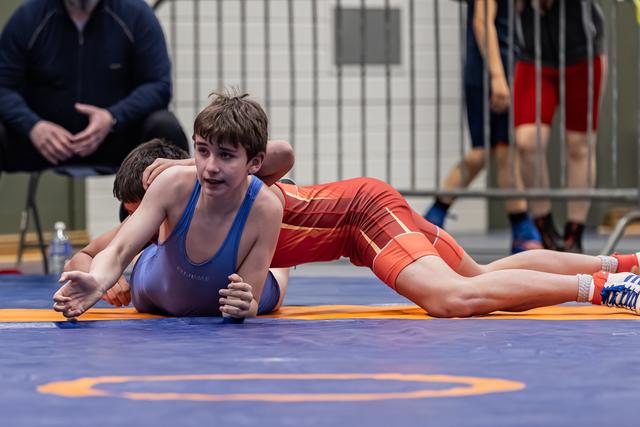 Young boy in blue singlet strains forward while opponent in red applies pressure from behind on a blue mat.