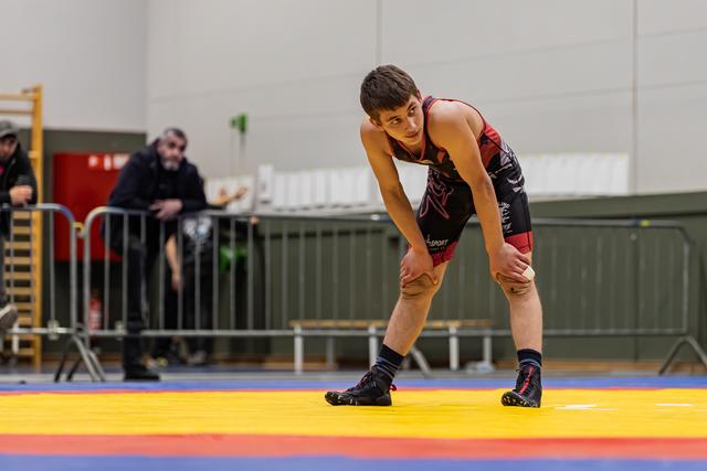 Teen wrestler in black and red singlet crouches in ready stance on colorful mat, looking focused and tired.