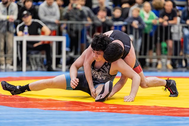 Young wrestler in black singlet resists opponent's takedown attempt, legs spread wide on a yellow and red mat.