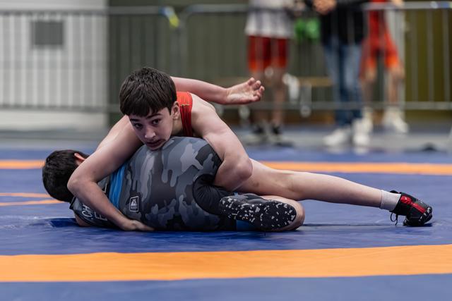 Teen wrestler in red singlet pins opponent on blue mat, focused expression, arm extended for balance.