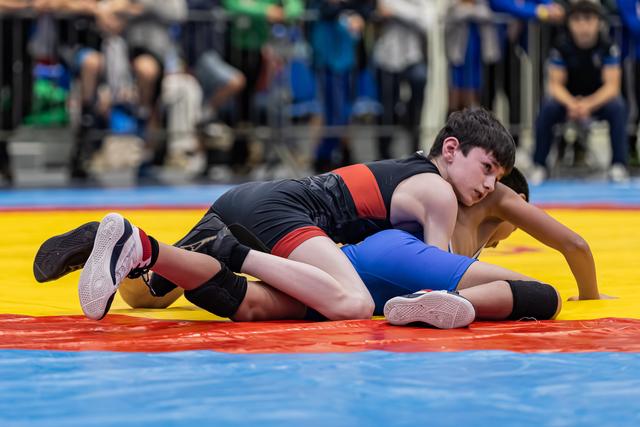 Young wrestler in black singlet controls opponent on the mat, focused expression during intense youth wrestling bout.