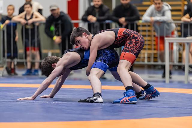 Teen wrestler in red-black singlet controls opponent on blue mat, spectators watching intently in background.