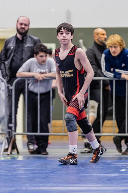 Young wrestler in black and red singlet stands alert on blue mat, focused expression, coaches watching behind barriers.