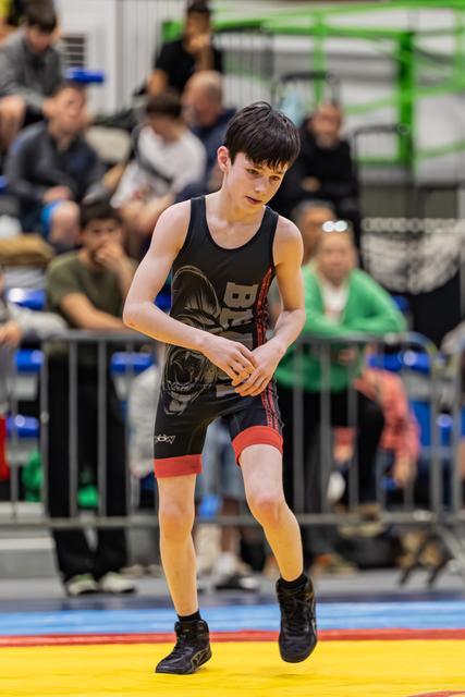 Young wrestler in black singlet moves with intense focus on colorful competition mat, crowd watching behind barriers.