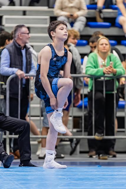Young male wrestler stretches his leg with focused intensity, standing on a blue mat before his match.