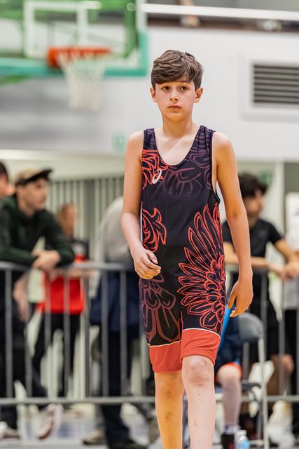 Young boy in ASICS singlet walks with calm focus across gymnasium floor during wrestling event.