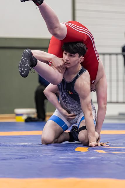 Focused teen wrestler lifts opponent overhead during match on blue mat inside sports hall.