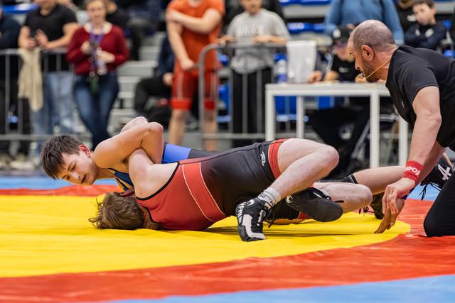 Young wrestler in red singlet pins opponent to colorful mat as referee leans in closely to observe the hold.