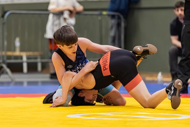 Young wrestler in blue singlet pins opponent to yellow mat, focused expression, indoor sports hall.