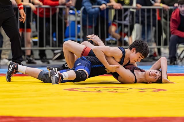 Young wrestler in blue singlet pins opponent to the mat, both showing intense focus during a youth match.