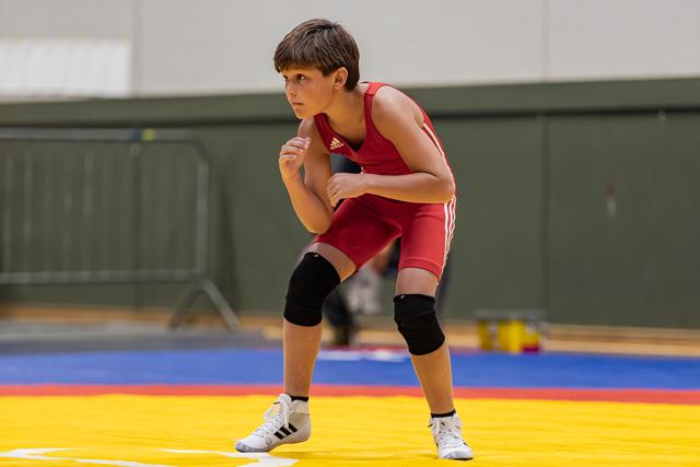 Young wrestler in red Adidas singlet crouches in fighting stance, focused and intense, on colorful competition mat.