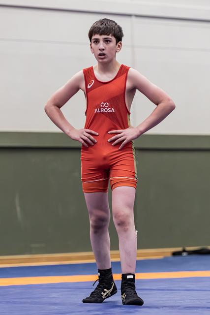 Young male wrestler in red Alrosa singlet stands hands on hips, alert expression, on blue competition mat.