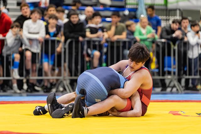 Wrestler in red singlet dominates opponent on yellow mat, crowd watching intently in background.