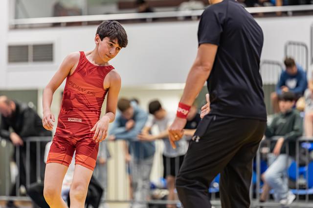 Young wrestler in red singlet crouches in focused stance, facing an adult coach on the mat during a sports event.