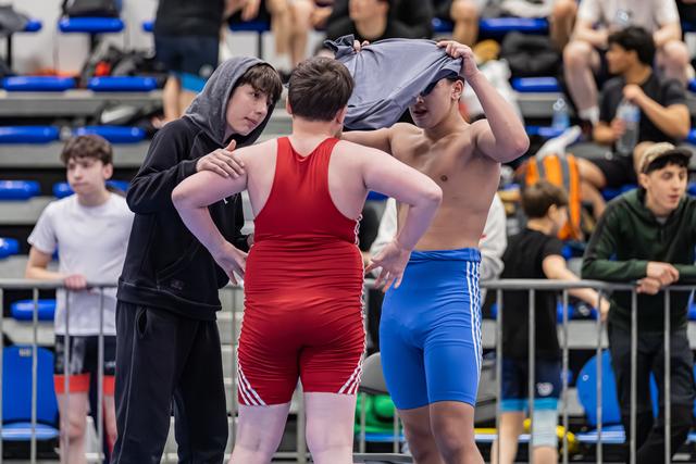 Teen wrestler in blue shorts removes shirt while a teammate in black hoodie offers advice before their bout.