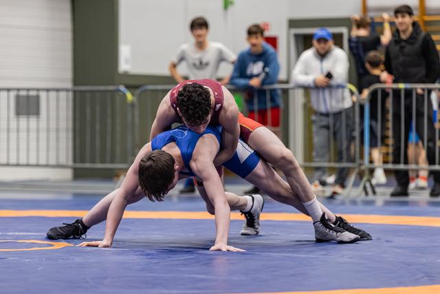 Wrestler in red singlet drives opponent in blue to the mat during an intense youth match indoors.