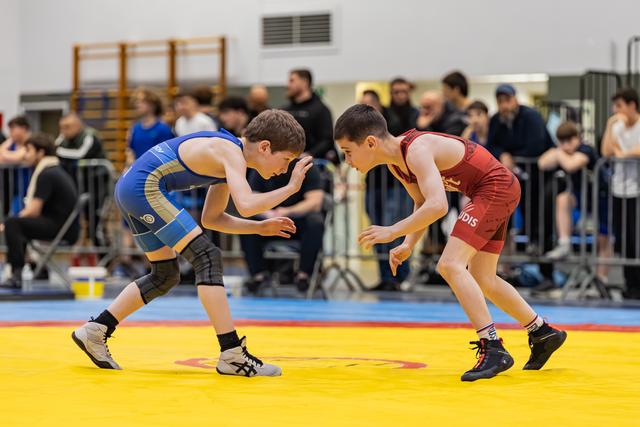 Two boys in blue and red singlets crouch in focused stance, circling each other on a yellow mat before a crowd.