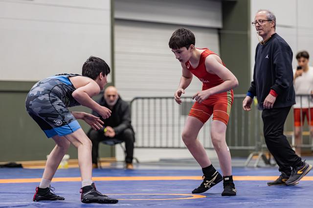 Two youth wrestlers in blue and red singlets crouch in tense standoff on blue mat, referee watching nearby.