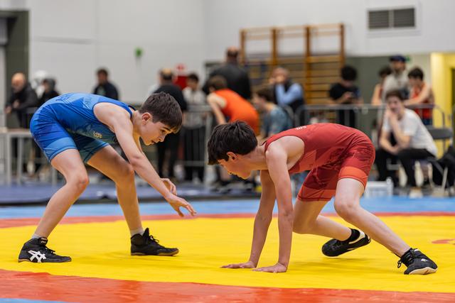 Two young male wrestlers in blue and red singlets crouch in tense readiness, eyes locked, on a yellow and red mat.