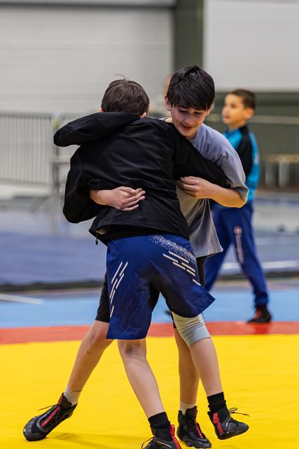 Two boys lock arms in a wrestling clinch on a yellow mat, one smiling, a third boy watches in the background.