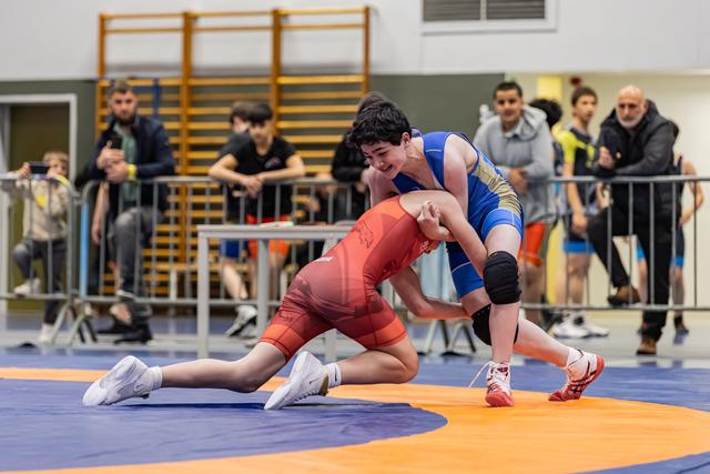 Two young wrestlers battle on the mat; blue singlet athlete drives forward aggressively while red singlet opponent braces low.