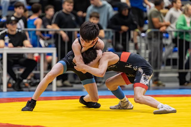 Two young wrestlers locked in a tight clinch, straining with effort on a colorful competition mat before a crowded arena.