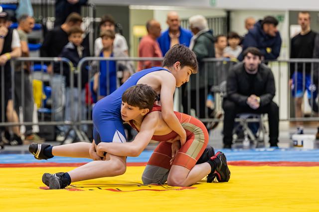 Two boys in blue and red singlets locked in an intense wrestling grapple on a yellow mat, crowd watching behind barriers.