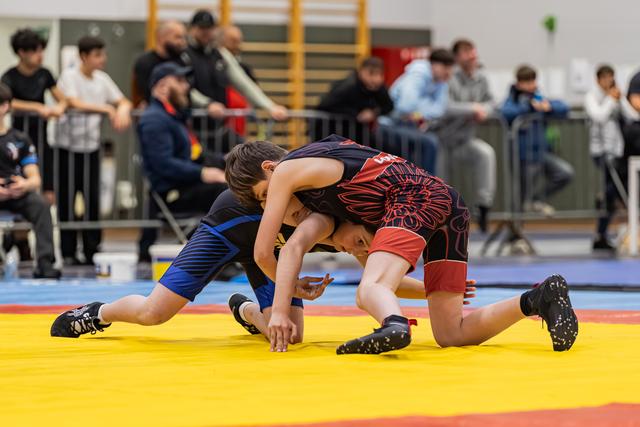 Two young wrestlers battle for control on the mat, one in blue pinned low by opponent in black and red singlet.