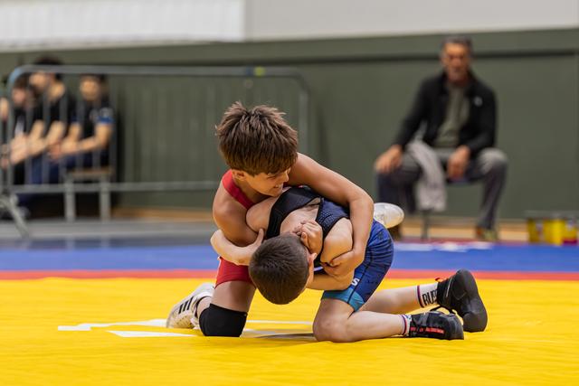 Two young boys wrestle on a yellow mat, one in red dominating with a headlock grip during an indoor tournament.