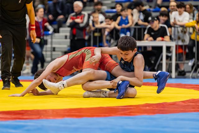 Two youth wrestlers battle on a colorful mat, one in red being controlled by opponent in blue grabbing his leg.